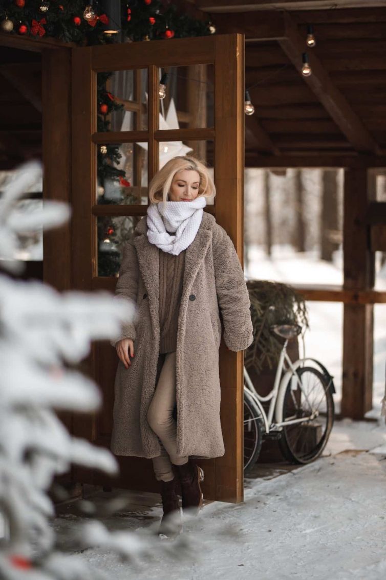 free-photo-of-blond-woman-wearing-a-beige-coat-and-white-scarf-standing-by-a-hut-with-christmas-decoration